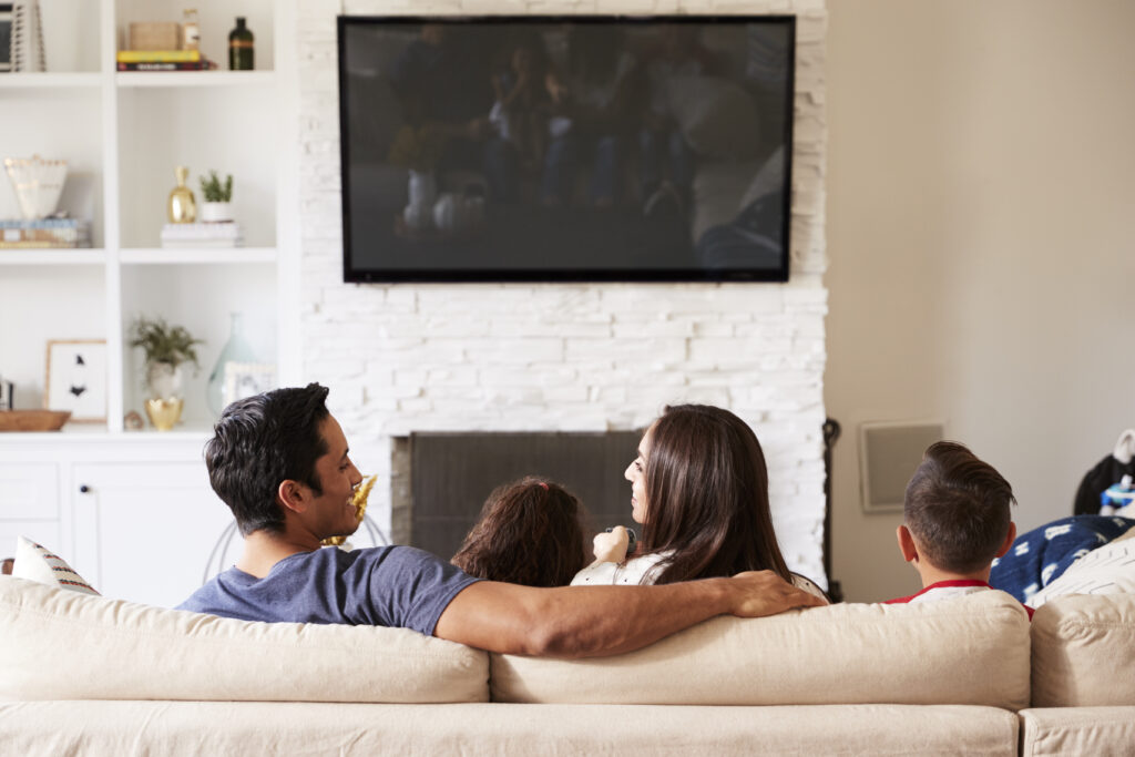 Back view of young family of four sitting on the sofa watching TV, mum looking at dad