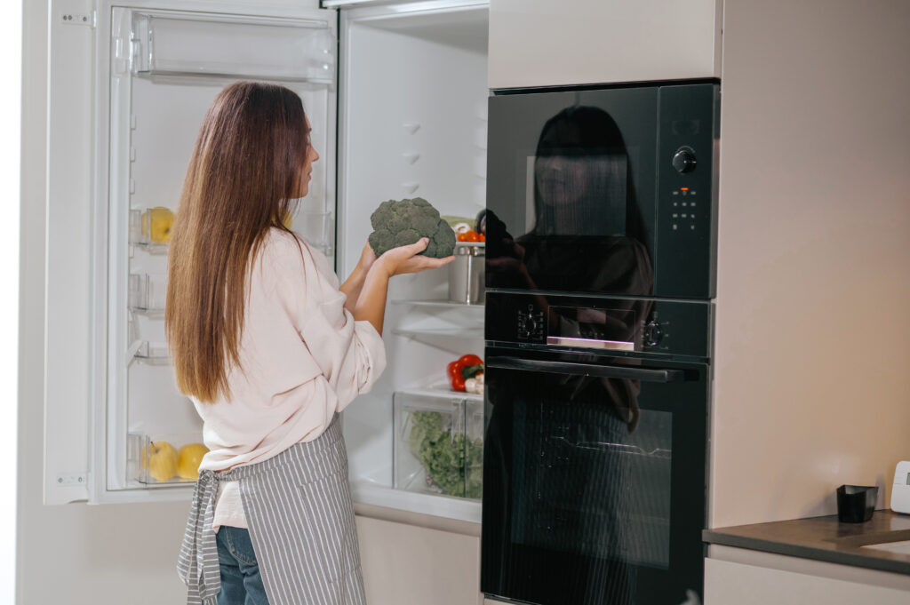 Woman holding broccoli while organising food in an open refrigerator.