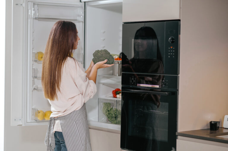 Woman holding broccoli while organising food in an open refrigerator.