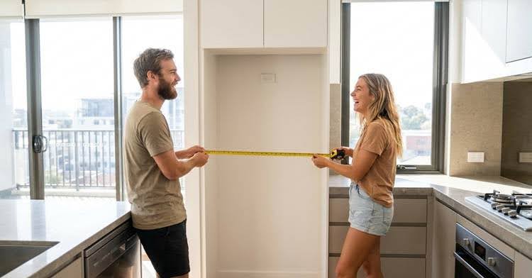 A smiling couple measures a kitchen space with a tape measure, planning for new appliances in their modern apartment.